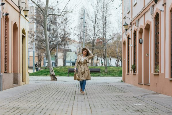 Front view of a woman wearing a coat and holding her beige hat on her head looking up walking between buildings in the city.