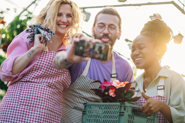 Portrait of a group of multiracial gardeners working at plant nursery and taking care of the flowers. Family business. Diverse gardeners working in a greenhouse and taking selfie with a smartphone.